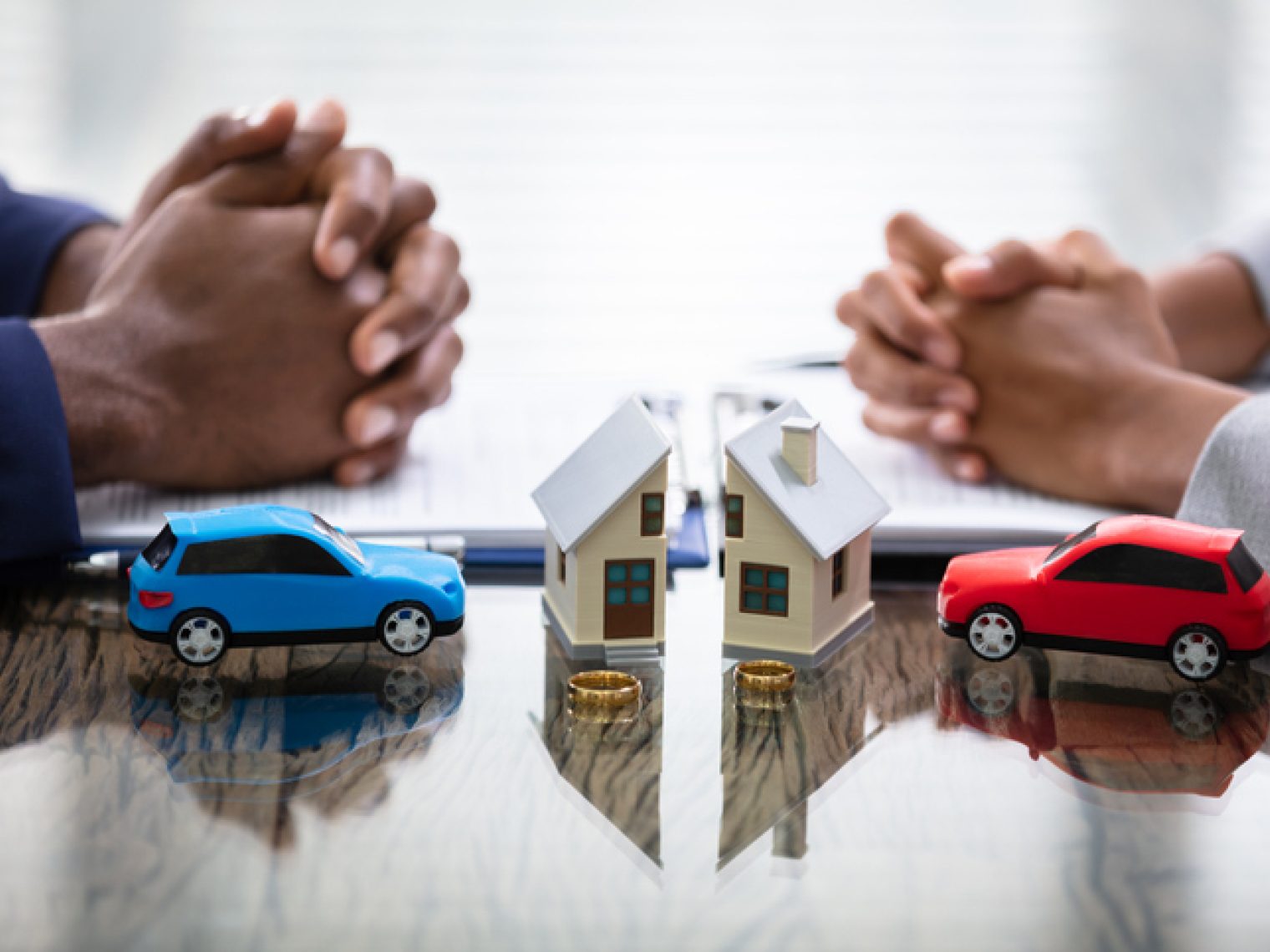 Two people with interlocked hands at a table, with toy cars and a small house model between them, symbolizing negotiations or discussions about property and automotive assets.