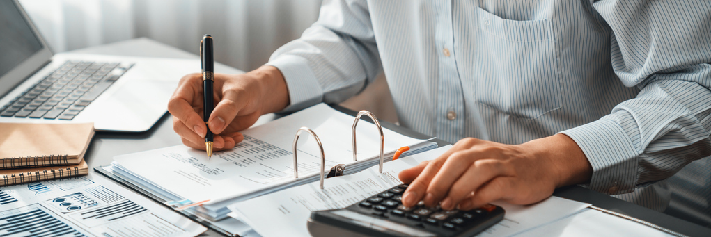 A person is working at a desk with financial documents, using a calculator with one hand and holding a pen in the other.