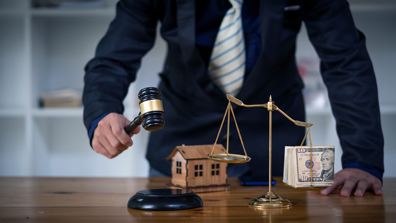 A person in a dark suit holds a judge's gavel in their right hand, positioned above a sounding block on a wooden table. To their side, there's a small model of a house and a balance scale with a stack of U.S. currency on one side.