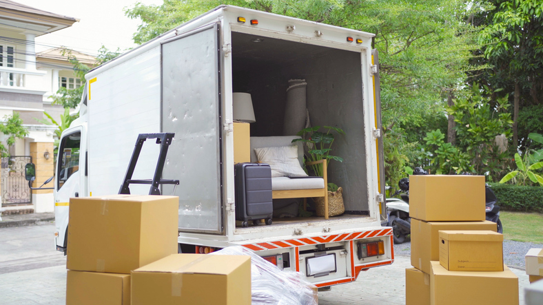 A moving truck is open and partially loaded with household items and furniture, with cardboard boxes stacked in the foreground, indicating a move in progress in a residential area.