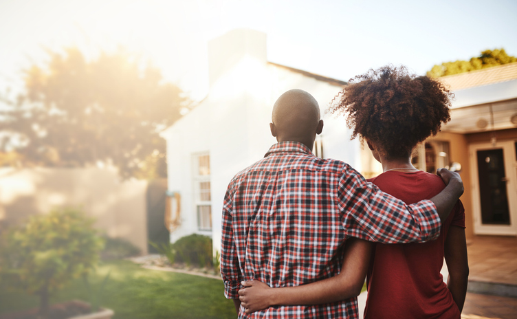 A back view of a couple embracing while looking towards a house with a sunlit garden.