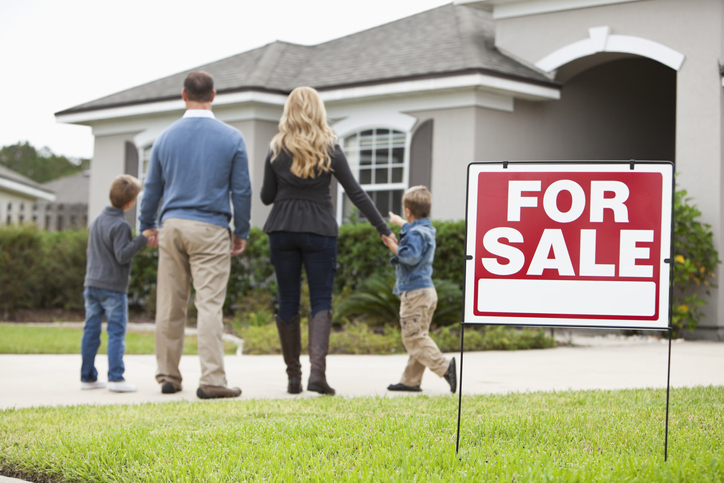 A family of four is seen from behind, walking towards a modern single-story house that is up for sale, indicated by a prominent red "FOR SALE" sign on the well-kept lawn.