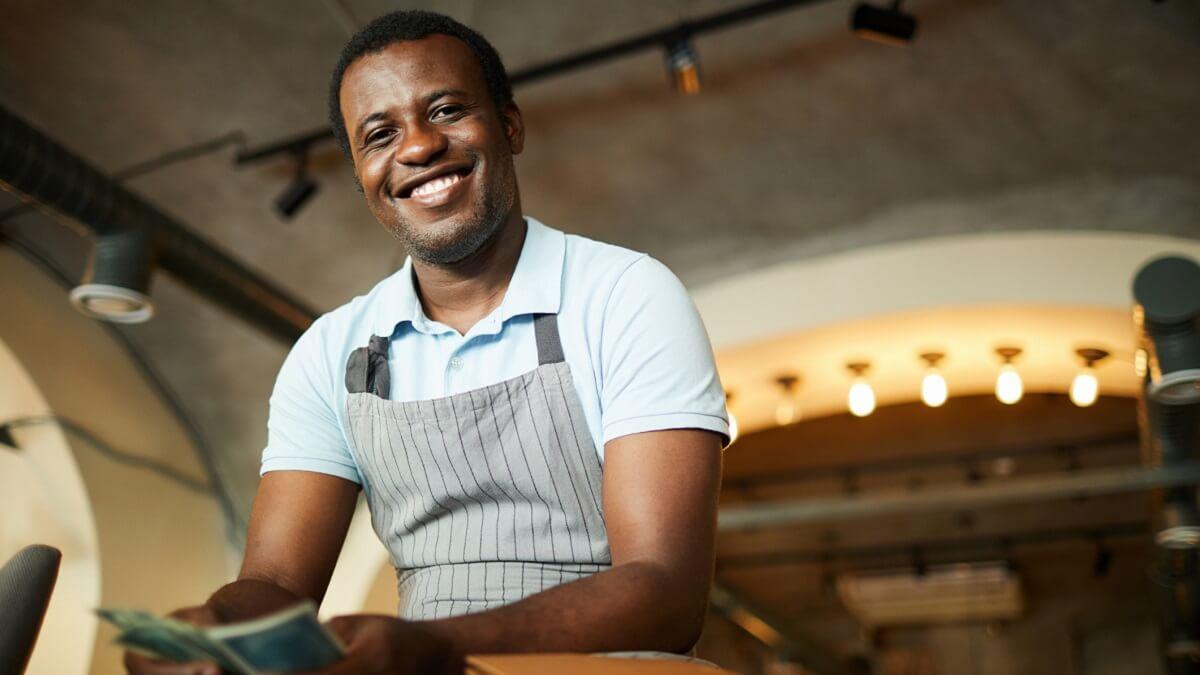 Man wearing an apron smiles at the camera while counting money.