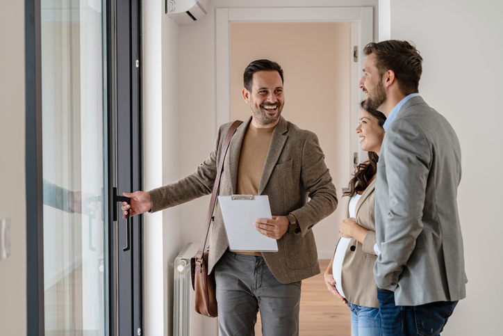 A real estate agent, holding documents, is smiling and gesturing welcomingly to a couple as they enter a home.