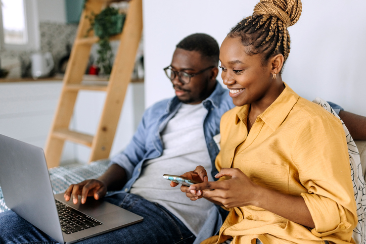 A woman is sitting next to a man who is focused on a laptop. Both are comfortably seated on a couch with pillows, suggesting a relaxed home environment.