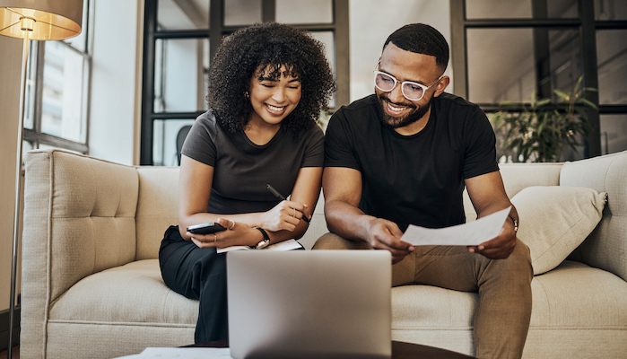 A couple sitting on a couch looking at a laptop smiling with pen and paper in hand.